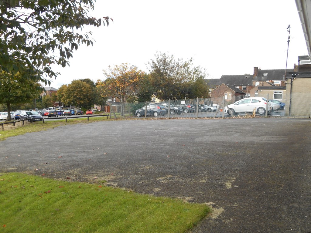Image shows tarmacked car park at rear of Meeting House. A public car park can be seen behind with a step over fence between the two.